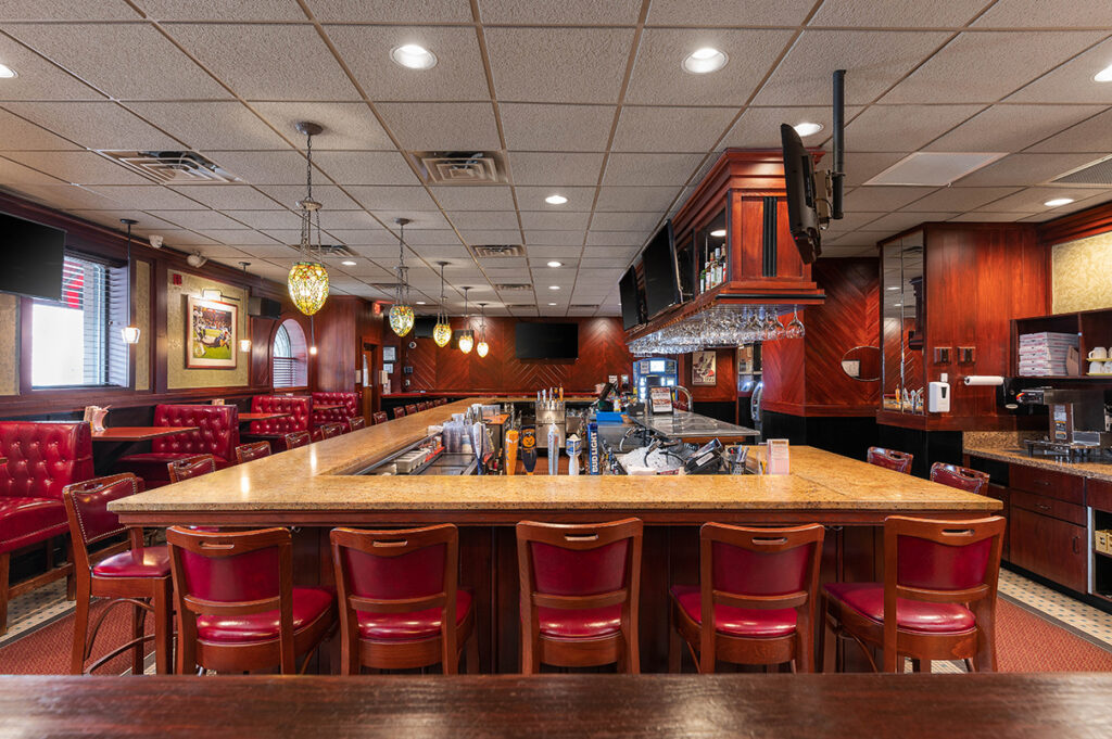 Classic restaurant bar interior with red leather stools, wooden finishes, pendant lights, and a polished granite counter.