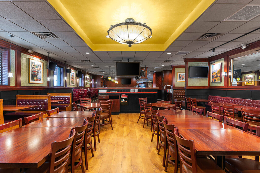 Empty restaurant dining area with wooden tables, red booth seating, warm lighting, and wall-mounted TVs.
