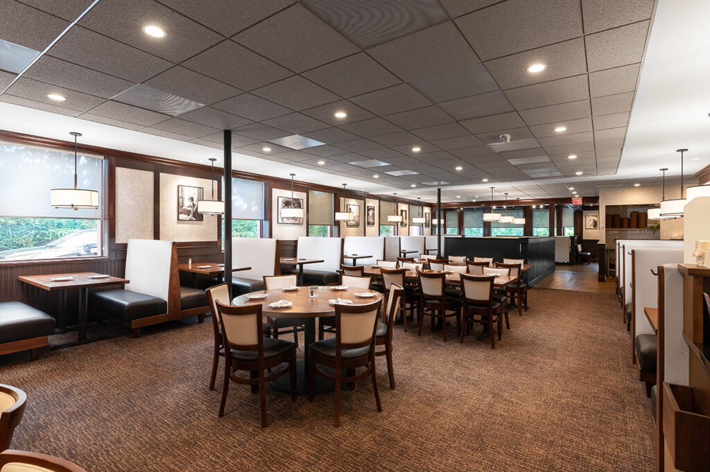 Bright restaurant dining room with booth seating, round tables, framed wall art, and warm overhead lighting.