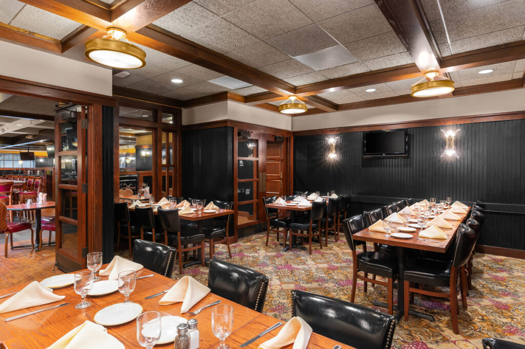 Cozy restaurant dining area with wood tables, black chairs, set place settings, and warm ceiling lighting.
