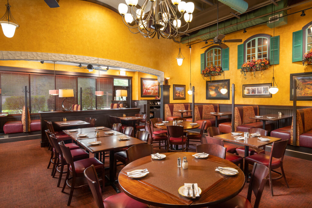 Warm Italian restaurant dining room with yellow walls, chandeliers, wood tables, and red upholstered booths.
