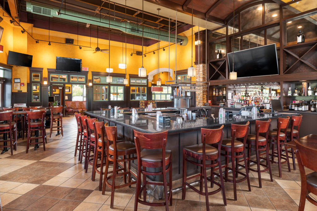 Spacious restaurant bar with granite countertop, wooden bar stools, hanging lights, and fully stocked back bar.