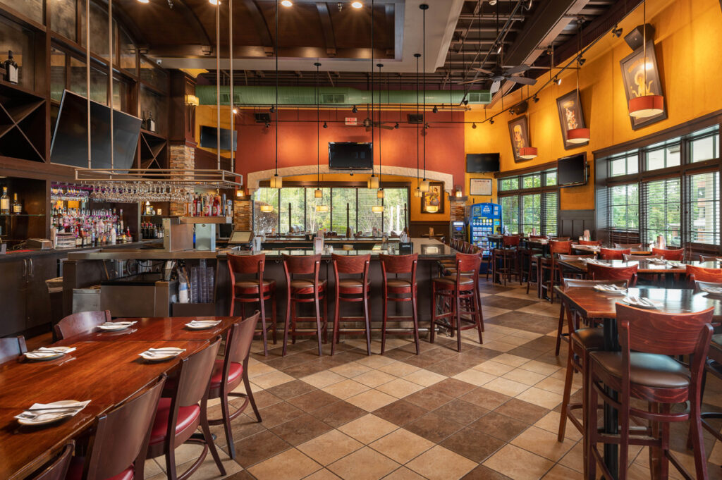 Wide view of restaurant dining area with central bar, wooden tables and chairs, warm lighting, and large windows.