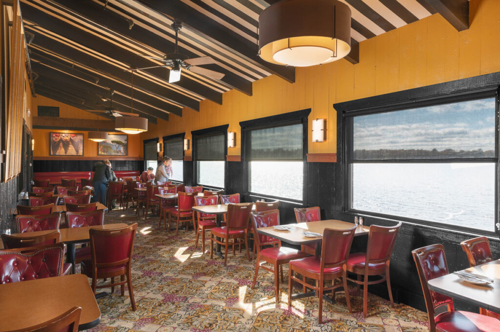 Bright dining room with red chairs and tables, large windows overlooking the water, and striped ceiling with warm lighting.