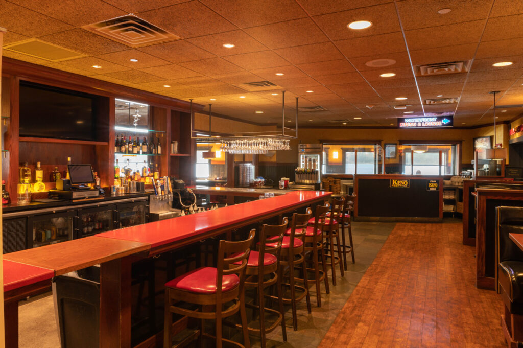 Restaurant bar area with red countertop, wooden stools, liquor shelves, and warm ambient lighting.