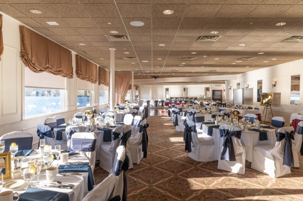 Banquet hall with round tables, white linens, navy chair sashes, and large windows overlooking the water.