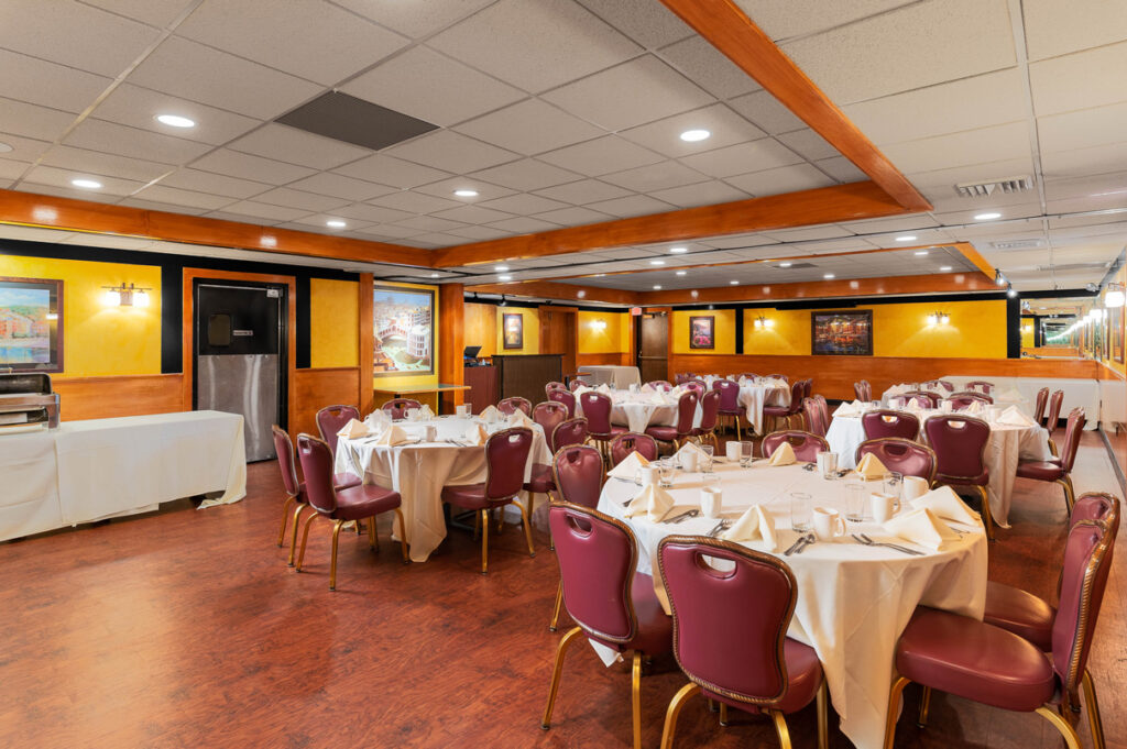 Banquet-style restaurant room with round tables, white tablecloths, burgundy chairs, warm lighting, and framed wall art.