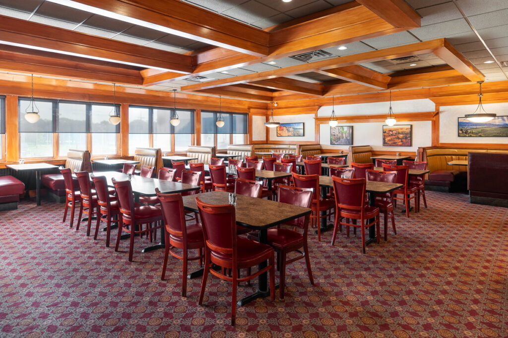 Spacious restaurant dining room with wooden beam ceiling, red chairs, booth seating, framed artwork, and large windows.