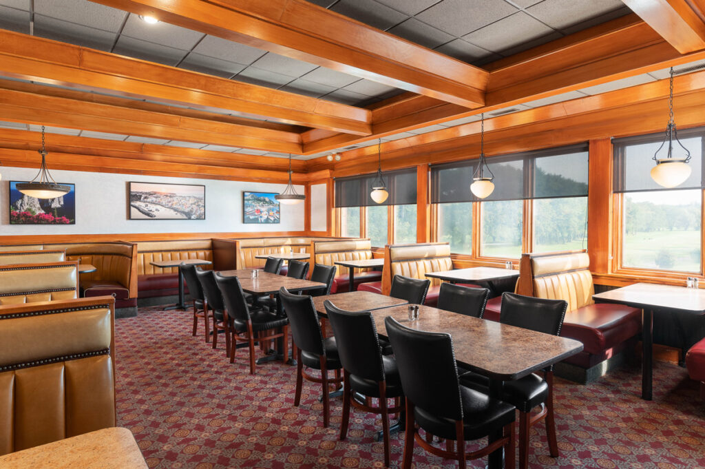 Cozy restaurant dining area with wooden ceiling beams, booth seating, tables and chairs, framed wall art, and large windows.