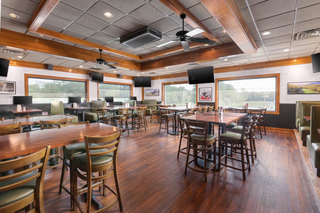 Bright restaurant dining room with wooden tables, high chairs, large windows, ceiling fans, and multiple wall-mounted TVs.