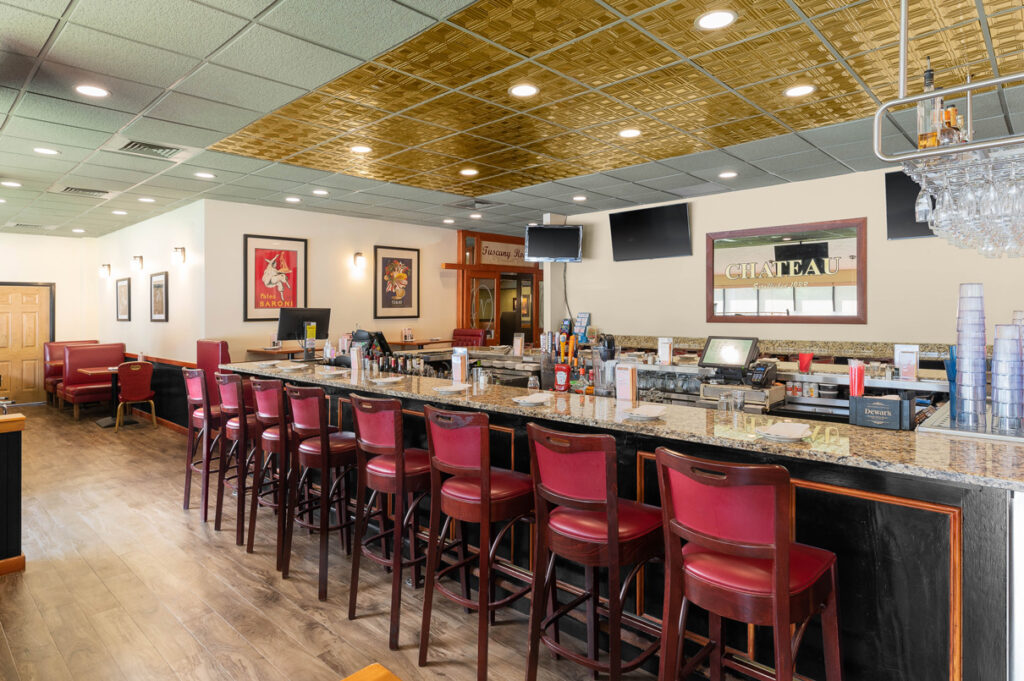 Interior bar area of The Chateau restaurant with granite countertop, red bar stools, hanging glassware, and warm lighting.