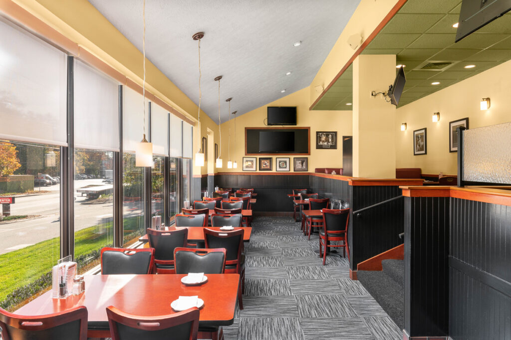 Bright dining area with window seating, wooden tables, black chairs, pendant lights, and TVs at The Chateau restaurant.