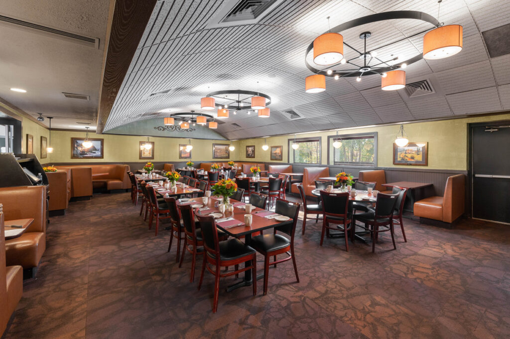 Spacious restaurant dining room with set tables, booths, floral centerpieces, and modern ceiling lights in a warm, inviting interior.